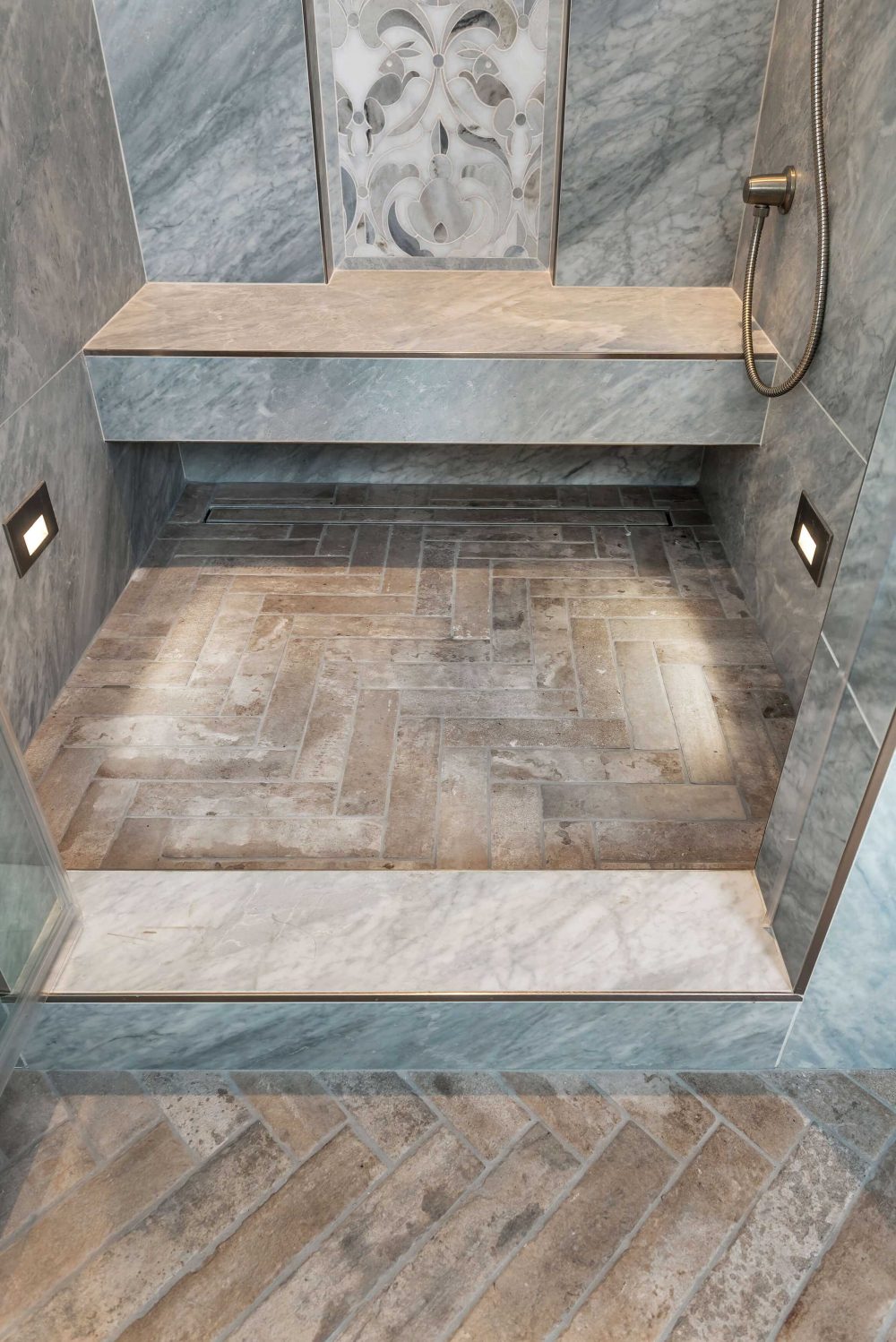 Close up of shower stall and patterned floor made from different stone. Built-in bench with grey and stone floral inlay design