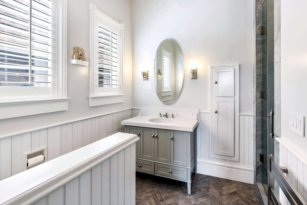White wainscoting bathroom showing sink, vanity mirror, built-in storage. Natural light through two windows.