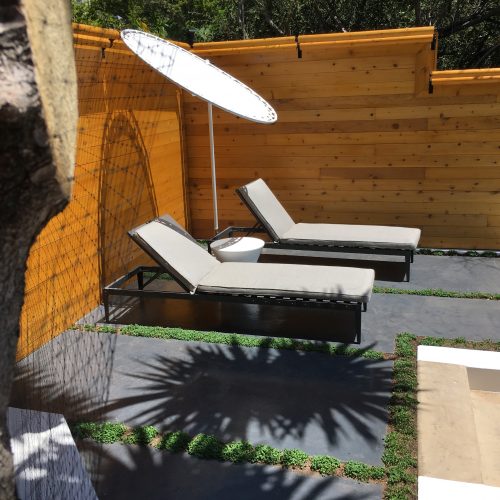 Small elevated patio next to fence with grey stained concrete slabs framed by grasses. Two lounge chairs and shade umbrella.
