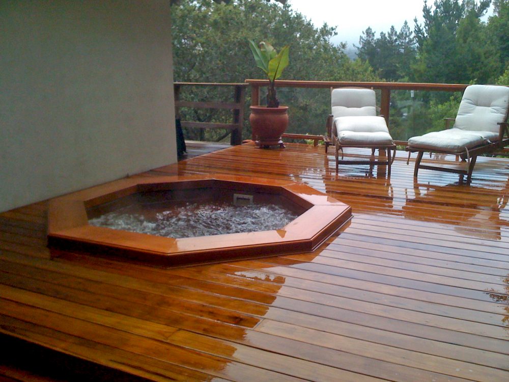 Large redwood deck around sunken hot tub next to side of house with plants and two cushioned lounge chairs