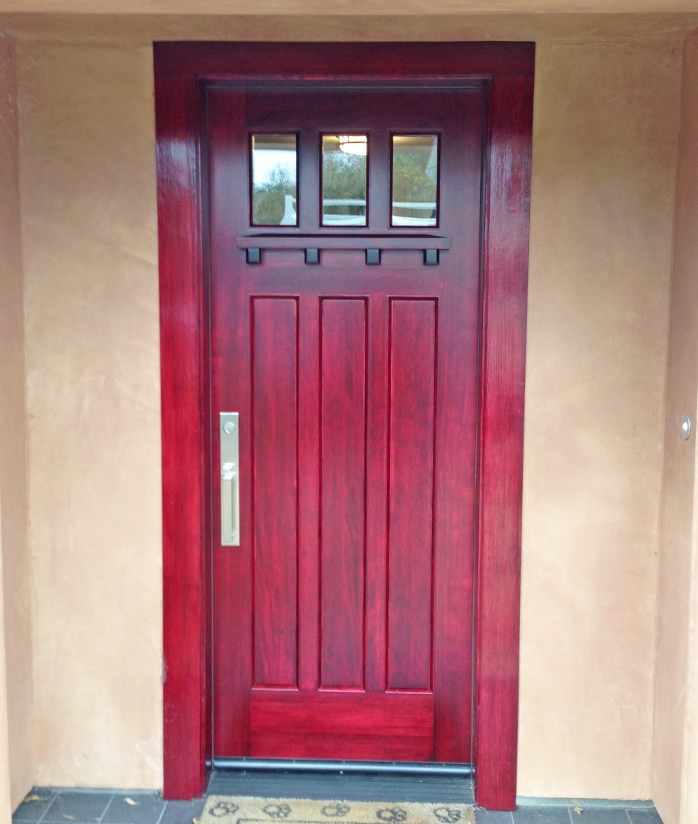 Three panel exterior door painted in deep red with 3 pane windows and wood molding under the windows; modern long silver colored door lock