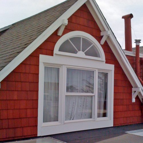 Large four pane gable window and arch window above it with white molding on a deep redwood colored shingled house