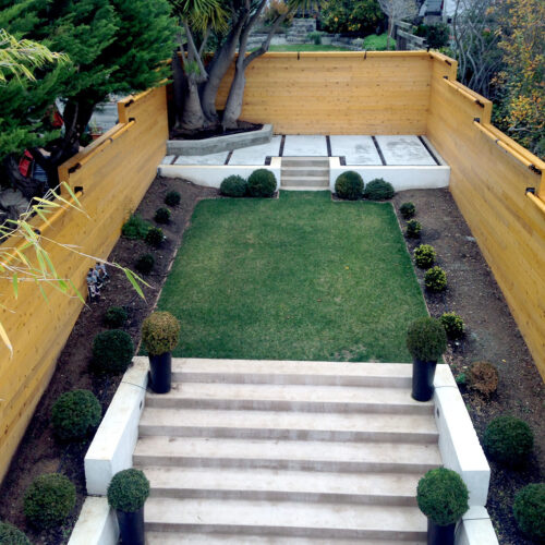 Arial view of stone steps leading up to a grassy area and third elevated cement patio surrounded by slat fencing and landscaping