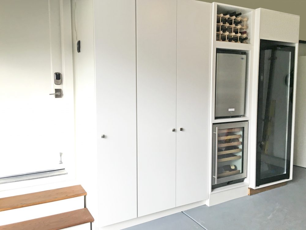 Very clean garage storage unit with three floor to ceiling enclosed cabinets next to the interior door, and wine storage coolers, half refrigerator, and vertical freezer units.